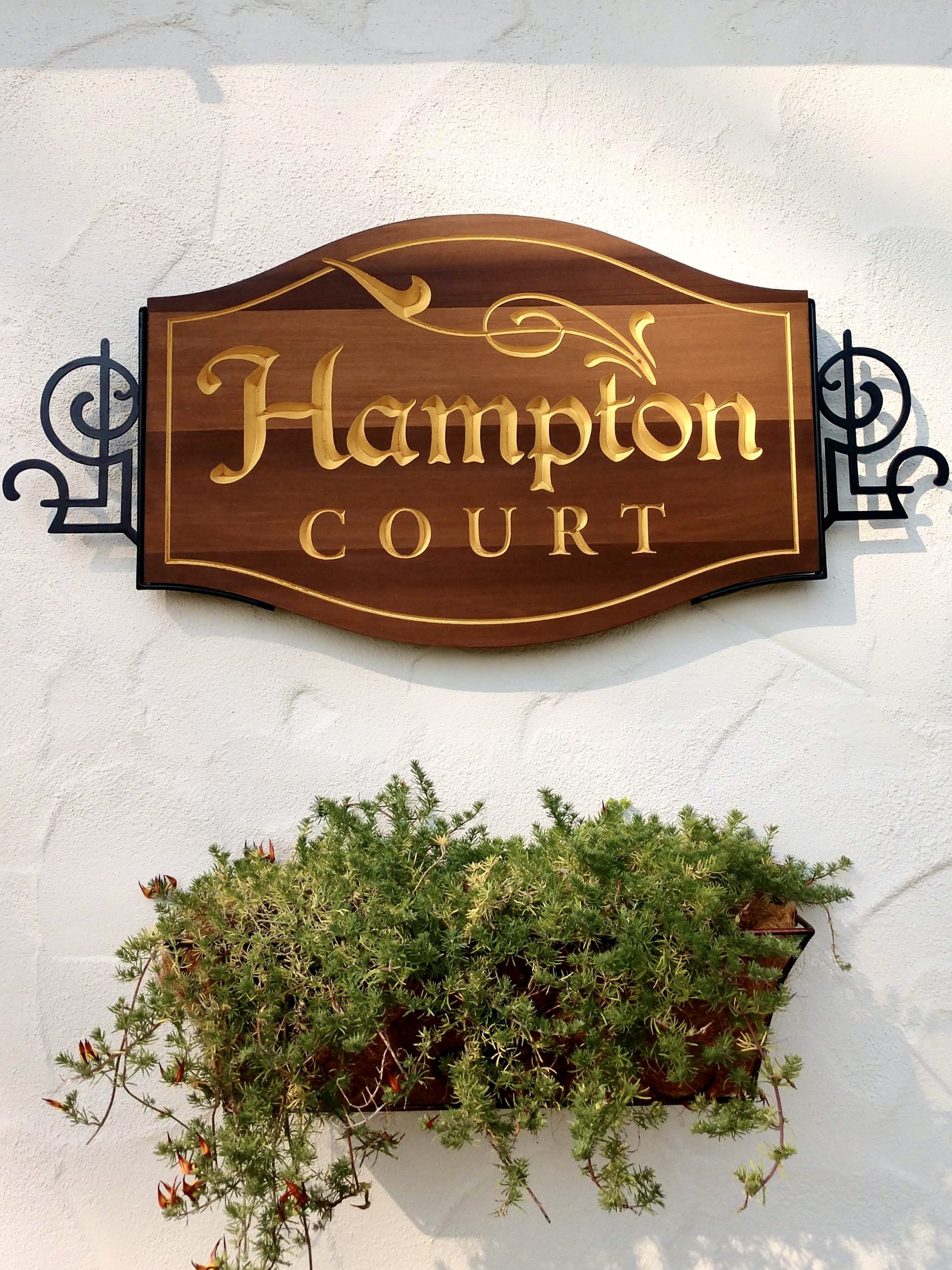 Close-up of Hampton Court wall sign showing routed cedar with metallic gold lettering and custom wrought iron scroll brackets